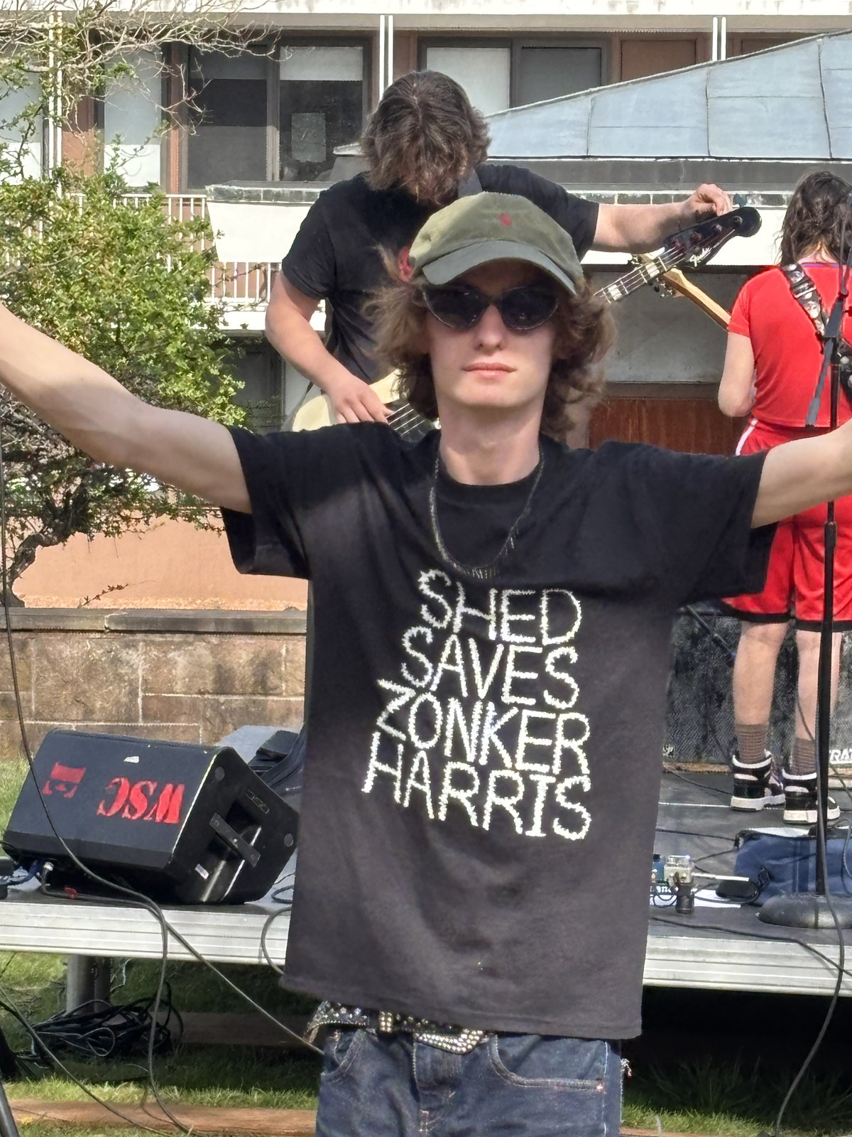 Ezra performing outdoors in a "Shed Saves Zonker Harris" t-shirt, arms out, bandmates playing bass and singing behind him.